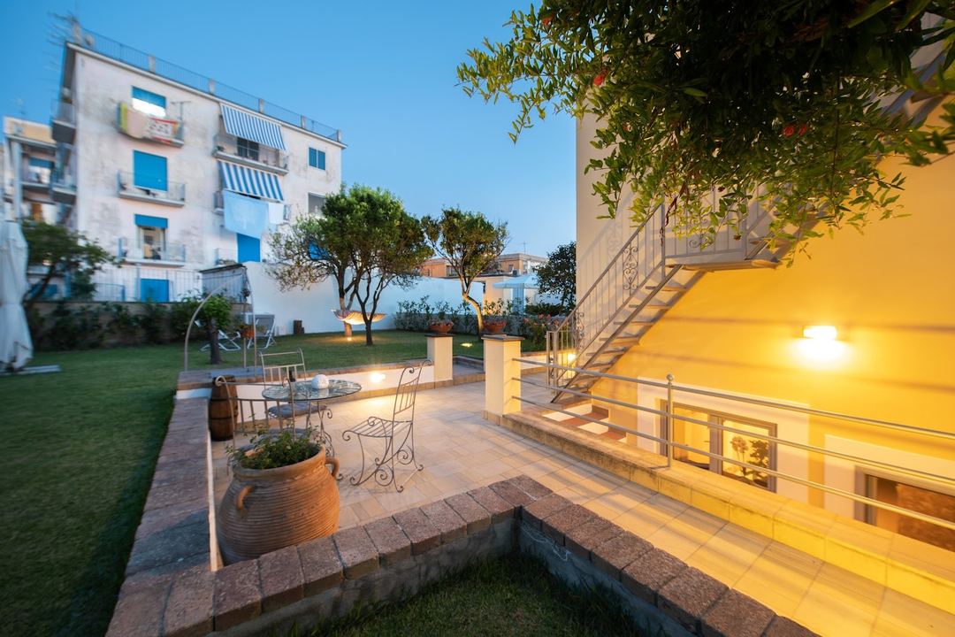 Twilight view of the B&B's courtyard terrace with illuminated yellow building facade, wrought iron seating, mature trees, and Mediterranean-style architecture.