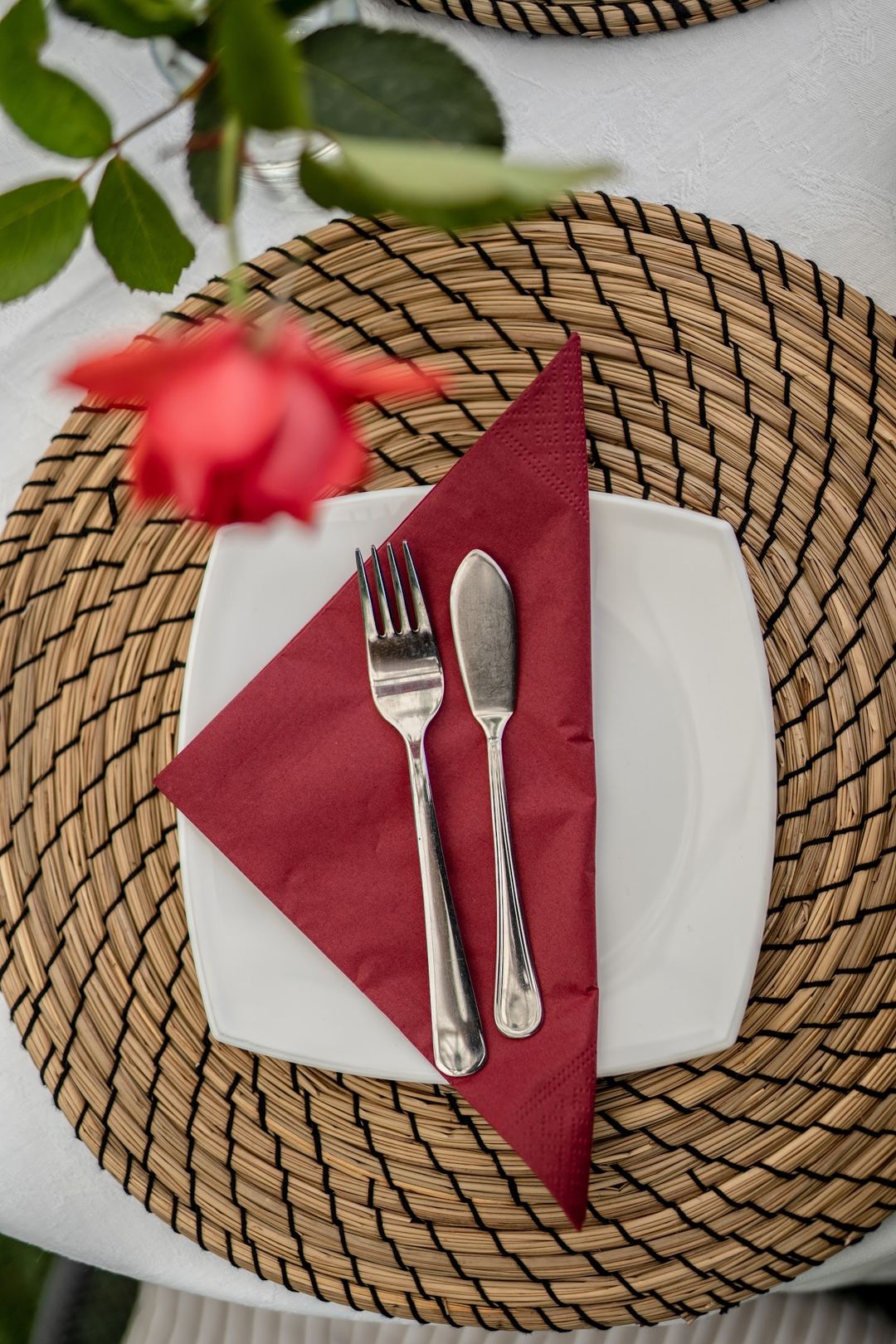 An elegantly plated table setting features a white plate with burgundy napkin, silver flatware, and woven placemat, garnished with fresh green leaves and a pink flower.