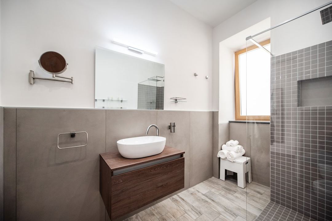 A modern bathroom features a white oval vessel sink on a dark wood vanity, gray tile accents, a walk-in shower with glass enclosure, and bright overhead lighting.