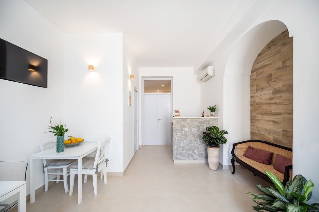 A bright, minimalist hotel room with white walls, wooden archway, dining area, and comfortable seating nook.
