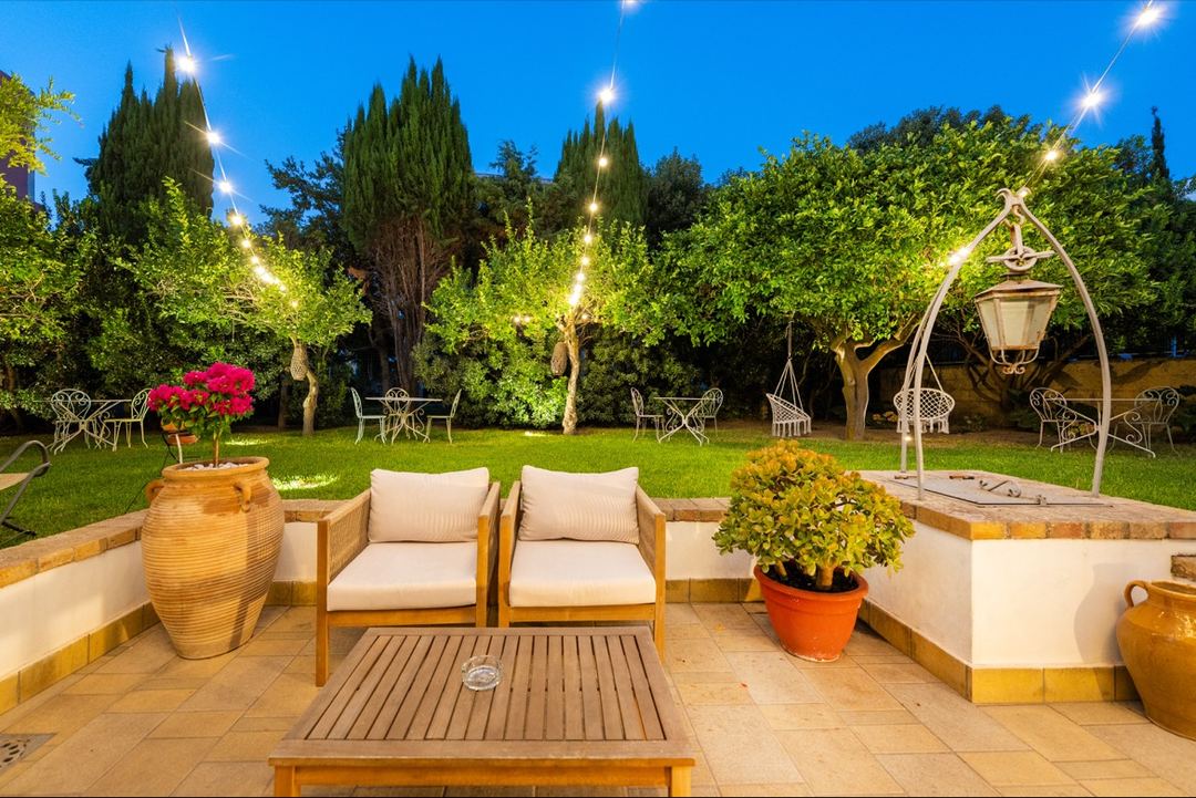 Evening view of an illuminated garden terrace with comfortable seating, potted plants, and cypress trees creating a Mediterranean ambiance.