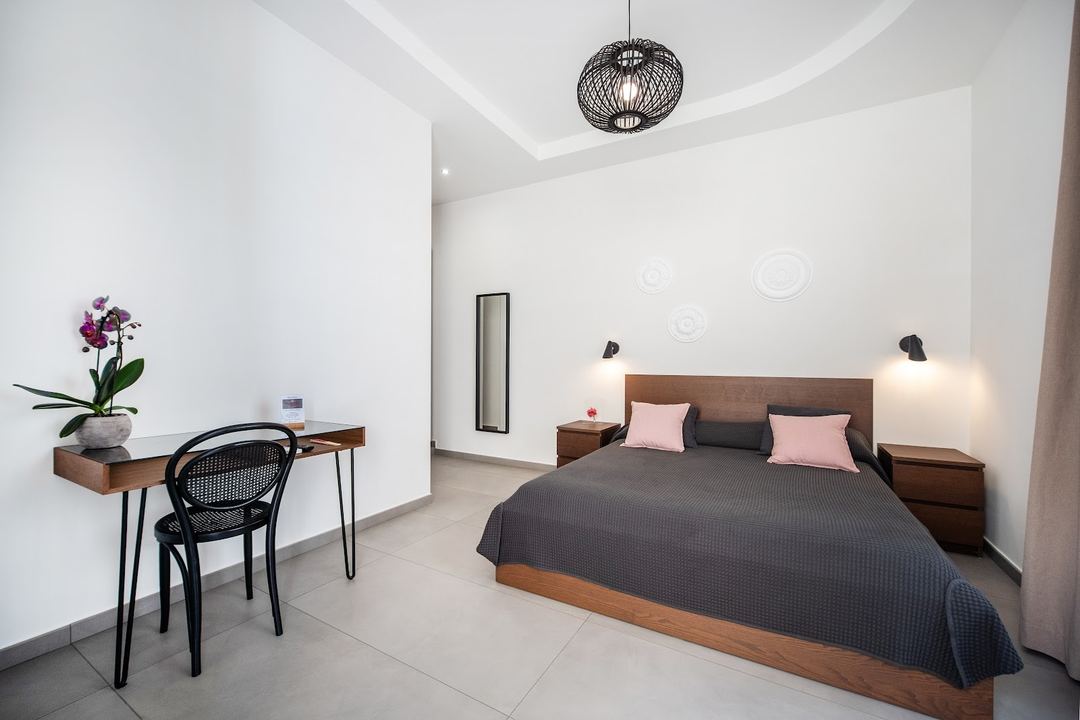A modern bedroom with a wooden bed frame, pink and charcoal bedding, desk workspace, and contemporary ceiling fixture in a minimalist design.