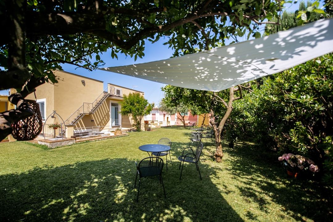 A sunny backyard garden area with seating under a shaded sail, featuring mature trees, manicured lawn, and a yellow stone building with terrace.