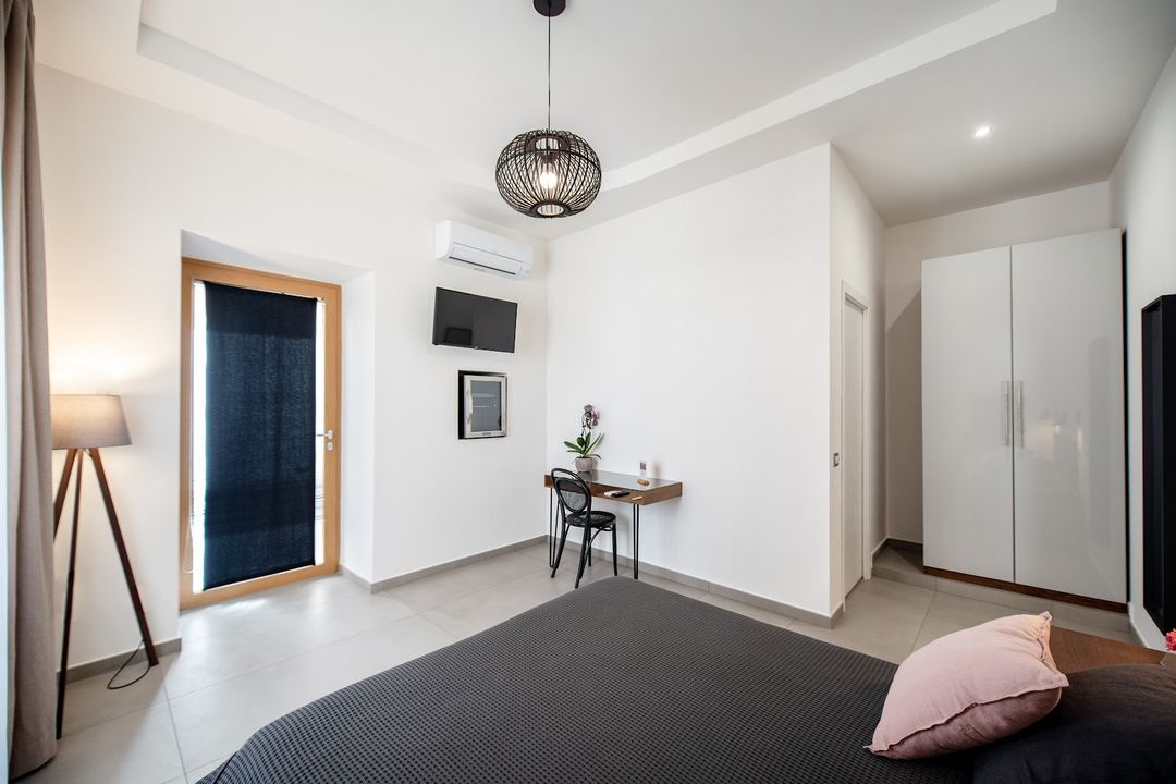 A modern bedroom with a dark bed, wooden desk workspace, white wardrobe, and pendant lighting in a minimalist design.
