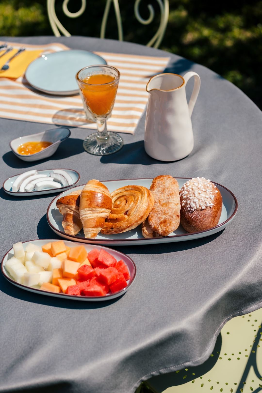A sunny outdoor breakfast spread featuring pastries, fresh fruit, orange juice, and cream pitcher served on a striped table at the B&B.