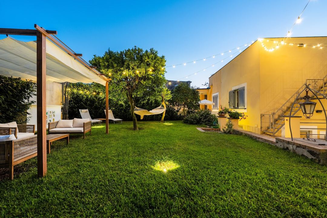 A beautifully landscaped evening garden with lounge seating under a pergola, string lights, and a yellow Mediterranean-style building.
